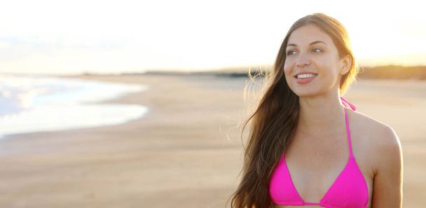 Νεαρή γυναίκα με ροζ μπικίνι απολαμβάνει το ηλιοβασίλεμα στην παραλία με τα μαλλιά στον άνεμο. Πανοραμική θέα του χαμογελαστού εφήβου στο ηλιοβασίλεμα στην παραλία. - Φωτογραφία, εικόνα