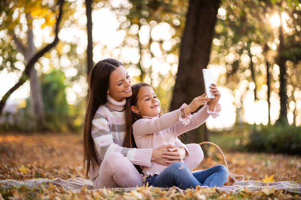 Matka i córka za pomocą smartfona jesienią w parku. - Zdjęcie, obraz