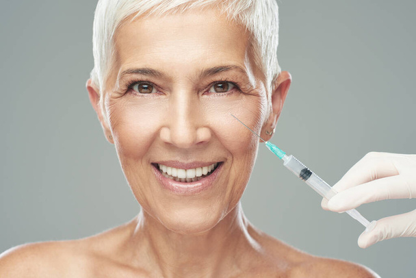 Beautiful smiling Caucasian senior woman standing in front of gray background and taking a shot of anti age product. Beauty photography. - Foto, afbeelding