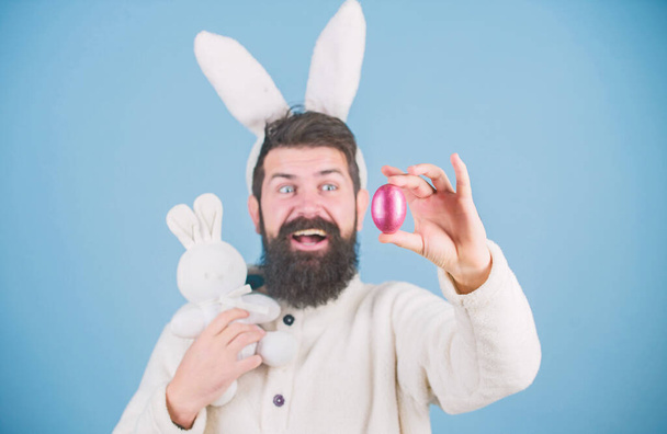 Decorating eggs for Easter. Bearded man with bunny toy and Easter egg. Hipster with long rabbit ears holding egg laying hare. Celebration of spring time holiday. Easter bunny delivering colored eggs - Zdjęcie, obraz