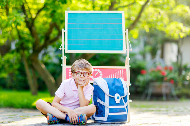 Ευτυχισμένος μικρό αγόρι παιδί με τα γυαλιά που κάθεται από το γραφείο και σακίδιο ή τσάντα. Schoolkid με παραδοσιακή γερμανική σχολική τσάντα καλεί την πρώτη του ημέρα στο σχολείο Schultuete. Γεια σας σχολείο στα Γερμανικά. - Φωτογραφία, εικόνα
