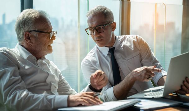 Two businessmen shaking hands together while sitting by windows.Mature businessman discuss information with a colleague in a modern business lounge high up in an office tower overlooking the city. - Foto, imagen
