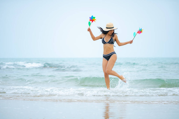 Sexy Asian woman in hat and black bikini jumping in water with windmills on beach. - Photo, image