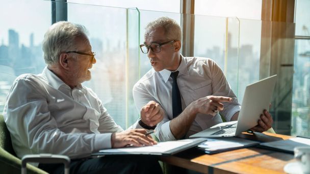 Two businessmen shaking hands together while sitting by windows.Mature businessman discuss information with a colleague in a modern business lounge high up in an office tower overlooking the city. - Valokuva, kuva