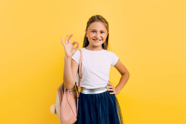 A little schoolgirl makes an approving gesture with her hand, OK on a yellow background. - Фото, зображення