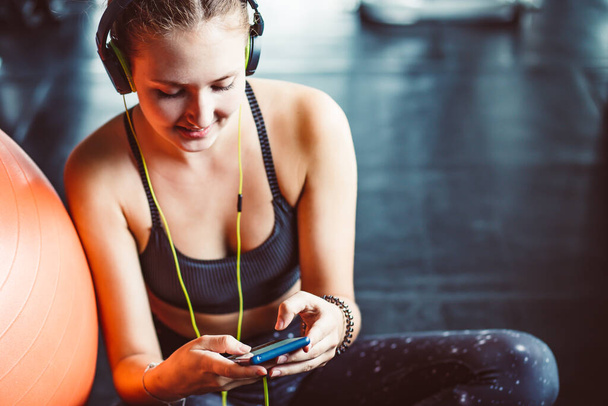 Young woman with earphones listening to music after hard workout in gym. Relax concept. Strength training and Body build up theme. Warm and cool tonePretty girl scrolling in her mobile phone. Close up - Zdjęcie, obraz