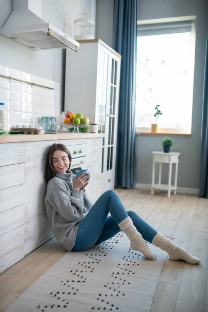 Dark-haired girl in a grey sweater sitting on the floor and having coffee - Foto, Bild