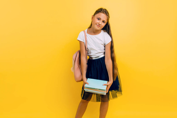 Tired teenage girl in school uniform holding a lot of books on an isolated yellow background - Foto, afbeelding