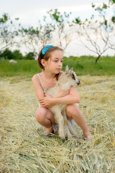 egy kiskecskés lány a szabadban. Falusi állatok. boldog gyermek ölelés kecske, koncepció a természet és az ember egysége. - Fotó, kép