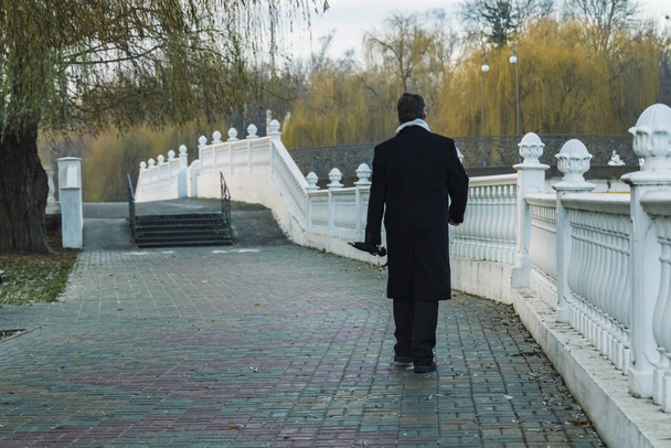 чоловік у чорному пальто повертається на роботу в парк на свіжому повітрі вранці холодний сезон погода з голими гілками дерев на асфальтовій доріжці з білою мармуровою стіною
 - Фото, зображення