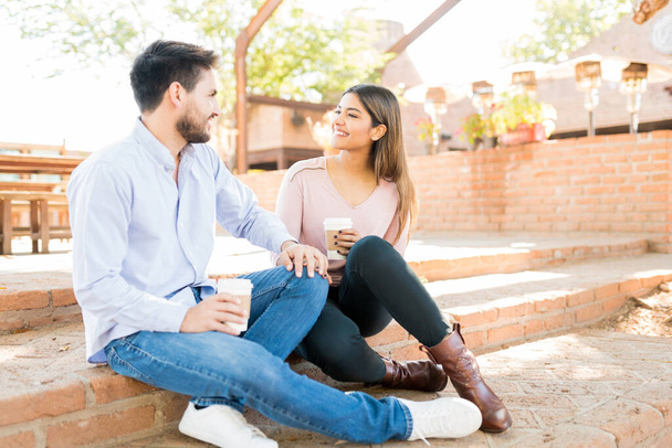 Sonriente novio latino y novia sosteniendo tazas de café desechables mientras está sentado en los pasos fuera de la cafetería
 - Foto, imagen