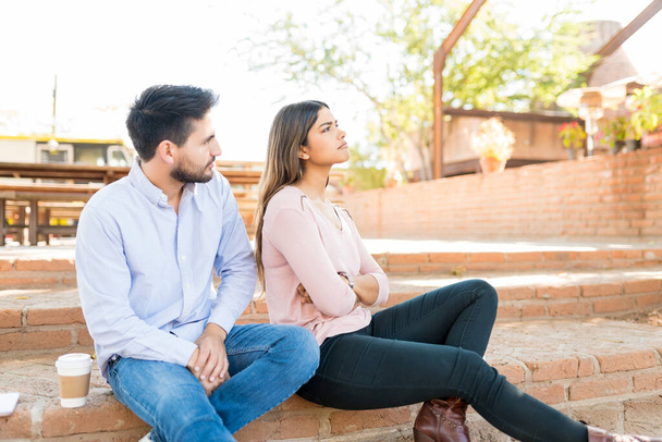 Ισπανόφωνος άντρας κοιτάζει απογοητευμένη κοπέλα ενώ κάθεται σε σκαλοπάτια έξω από το καφέ - Φωτογραφία, εικόνα
