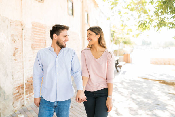 Happy Hispanic lovers holding hands while walking on footpath during sunny day - Photo, Image