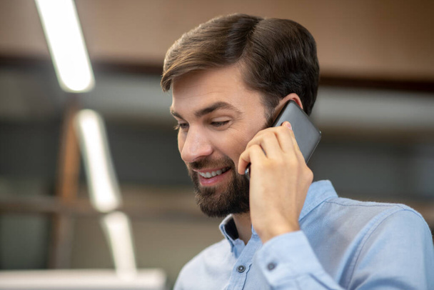 Young bearded man in a blue shirt talking on the phone and smiling - Foto, afbeelding