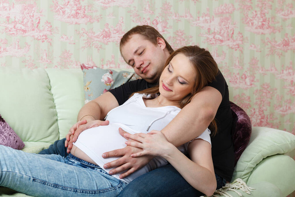 The happy pregnant woman with the husband sitting on a sofa of the house. - Photo, Image