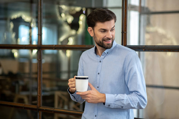 Young bearded man in a blue shirt smiling and having coffee - Fotó, kép