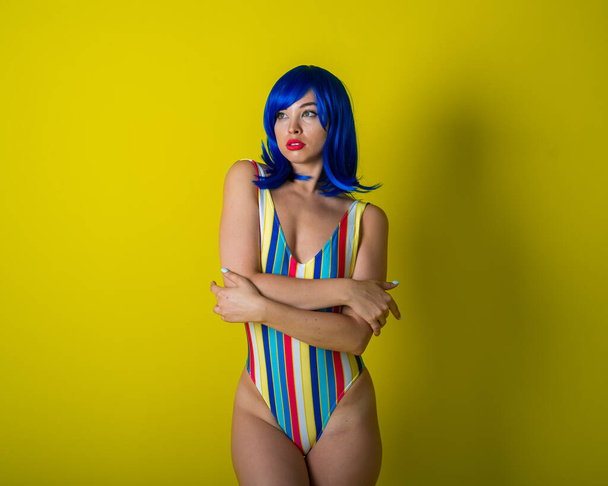 Beautiful woman in a striped swimsuit in a blue wig posing on a yellow background. Portrait of a glamorous girl. - Photo, Image