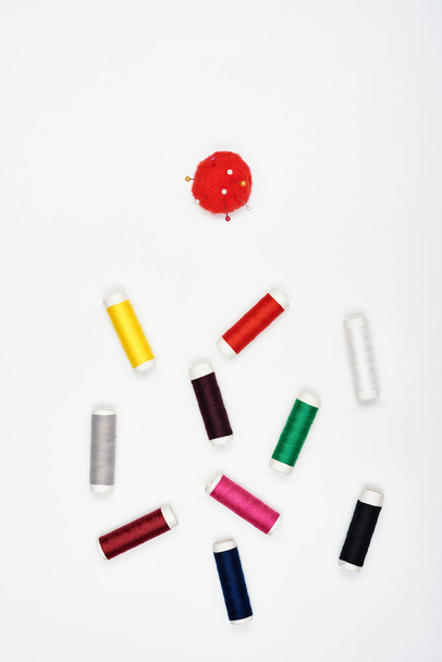 Bobinas con hermosos hilos multicolores para coser y un cojín rojo con alfileres en una superficie blanca. Vista superior
. - Foto, Imagen