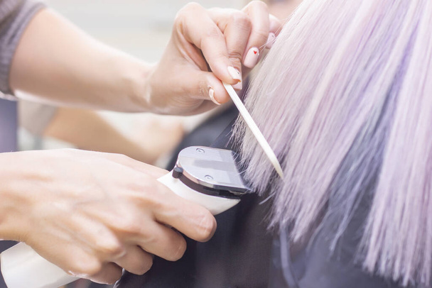 hairdresser makes haircut to a girl in with long blonde hair. Hairdresser hands holding trimmer and comb - Foto, imagen