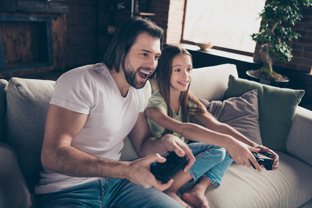 Photo of little pretty lady handsome dad sitting cosy sofa good mood holding joystick playing video games excited rejoicing one team work house room indoors - Photo, Image