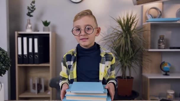 Portrait of good-looking 10-aged serious boy in glasses which holding a lot of books on table and looking at camera - Video