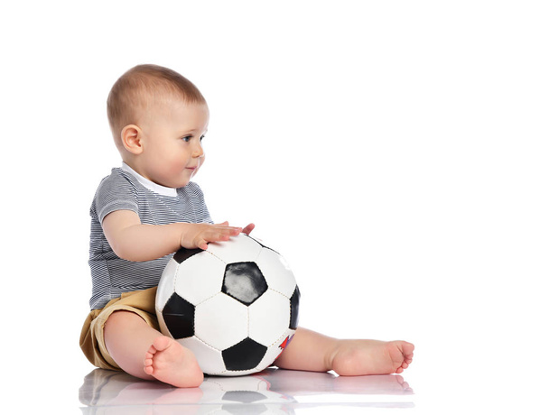 Infant baby boy toddler in striped t-shirt and pants is sitting holding football soccer ball playing on white - Foto, immagini