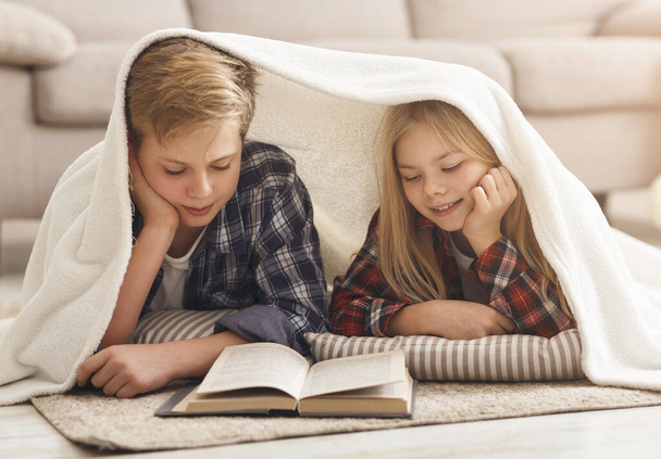 Hermano y hermana leyendo libro cubierto con manta en casa
 - Foto, Imagen