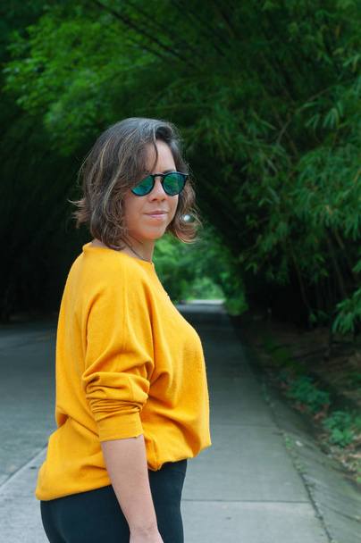 Kobieta w żółtym swetrze i okularach przeciwsłonecznych, z bezpośrednim spojrzeniem, na ścieżce bambusowej - Zdjęcie, obraz