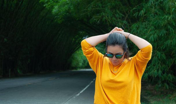женщина в желтом свитере и солнечных очках, делая хвостик, когда она идет по естественной бамбуковой дорожке в великий весенний день
 - Фото, изображение