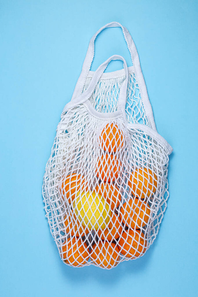 A white cotton string bag with tangerines and lemon lies on a blue background. - Fotografie, Obrázek