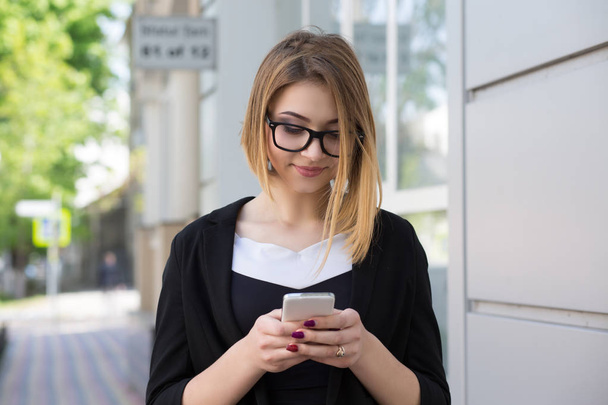 Singola signora felice utilizzando uno smartphone guardando on line contenuti in piedi sulla strada sorridente in abiti da lavoro e occhiali neri
. - Foto, immagini