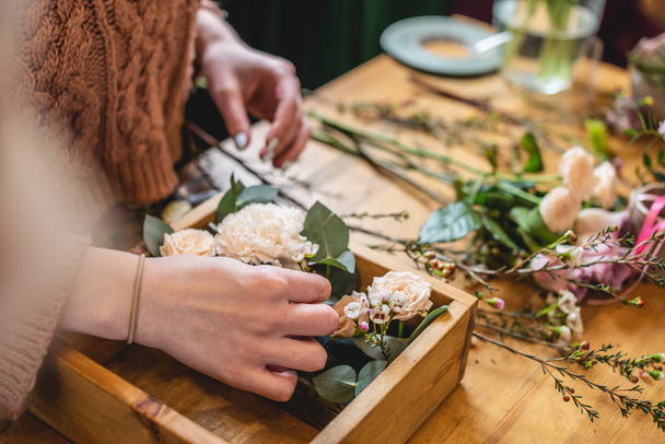 Γυναίκα ανθοπώλης συλλέγει μια σύνθεση λουλουδιών σε ένα ξύλινο κουτί. Άνοιξη μπουκέτο τριαντάφυλλα σε παστέλ χρώματα σε ένα ανθοπωλείο - Φωτογραφία, εικόνα