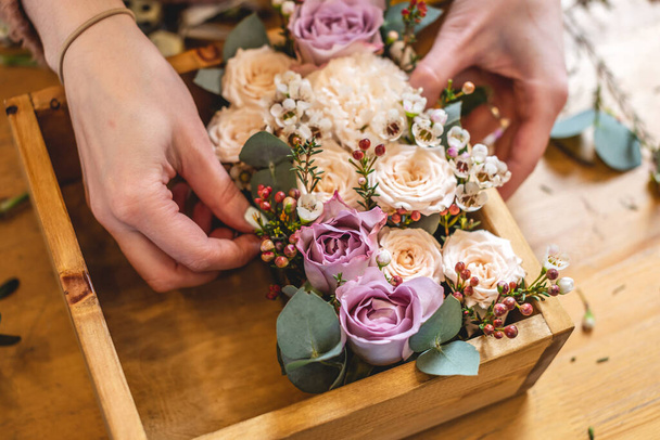 Mujer florista recoge una composición de flores en una caja de madera. Ramo de primavera de rosas en colores pastel en una florería
 - Foto, Imagen