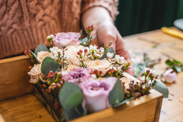 Woman florist collects a flower composition in a wooden box. Spring bouquet of roses in pastel colors in a flower shop - Foto, afbeelding