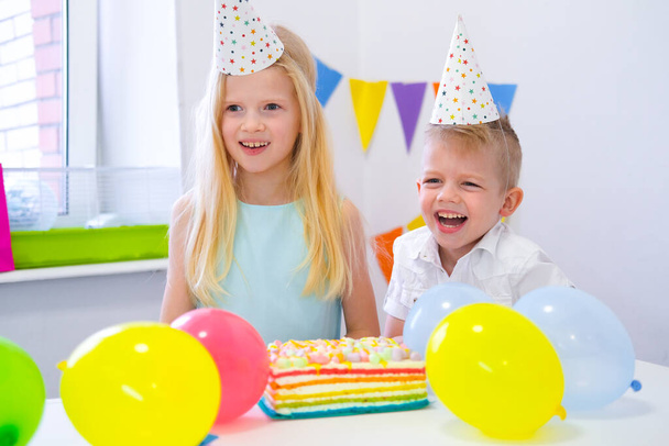 Dva blonďatí běloši kluci a holka se baví a smějí se na narozeninové oslavě. Barevné pozadí s balónky a narozeninový duhový dort. - Fotografie, Obrázek
