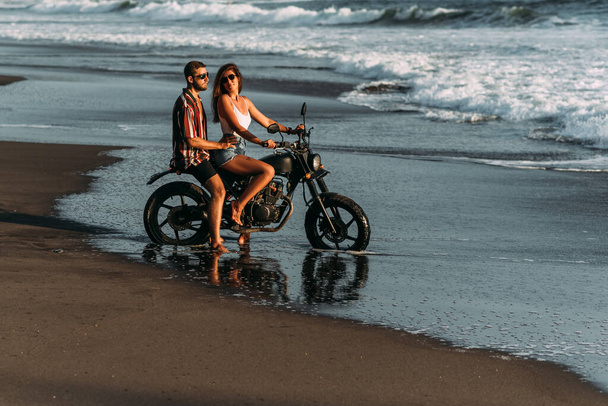Couple on a motorcycle on the beach. A couple in love on the beach meets the sunset. A man embraces a woman on the beach. Beautiful couple on a motorcycle. Travel on a motorcycle. Lovers on the beach - Foto, Bild