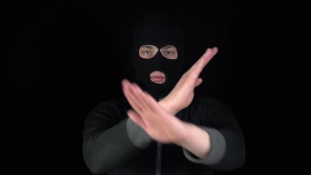 Ein maskierter Mann mit Sturmhaube zeigt mit seinen Händen ein negatives Zeichen. Der Schläger verschränkte seine Arme auf schwarzem Hintergrund. - Filmmaterial, Video