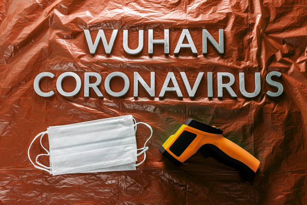 slova wuhan coronavirus položil s kovovými písmeny na oranžové zmačkané plastové fólie pozadí s obličejovými maskami a teploměrem - Fotografie, Obrázek