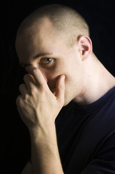 side head-and-shoulder portrait of a bald-headed young man with dark blue t-shirt against black background holding his hand to his forehead and looking embarrassed into the camera - Foto, imagen