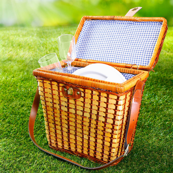 Fitted wicker picnic basket or hamper standing on fresh lush green grass with the lid open displaying a pretty blue and white checked lining with plates and glasses - Photo, image