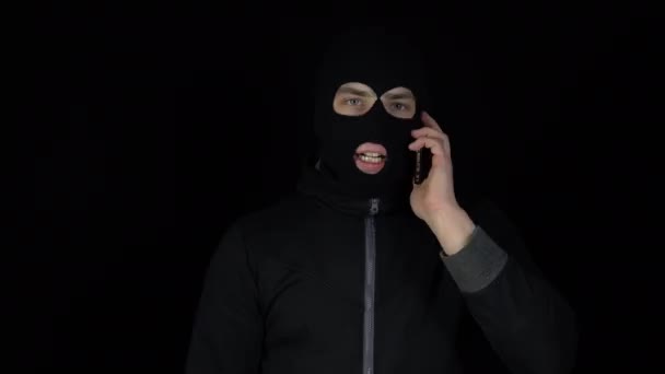 Un uomo con una maschera di passamontagna sta parlando al telefono. Un delinquente che urla al telefono su uno sfondo nero. - Filmati, video