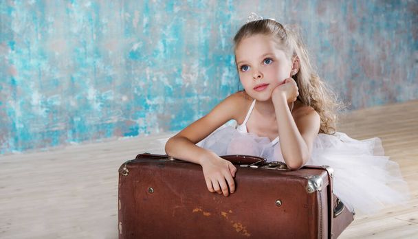 Little adorable ballerina in white tutu with old vintage suitcase in a beautiful studio. Travel and voyage concept. - Photo, Image