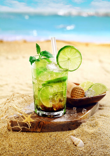 Caipirinha Rum and lime cocktail garnished with mint leaves and served on a wooden board on the golden sand of a tropical beach - Valokuva, kuva