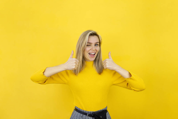 Smiling blonde young woman in yellow showing two thumbs up and looking in camera. Studio shot on yellow background - Φωτογραφία, εικόνα