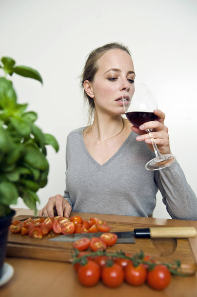 Attractive young housewife enjoying drinking a glass of red wine while standing in the kitchen cooking and slicing tomatoes - Φωτογραφία, εικόνα