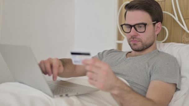 Joven barba con éxito utilizando tarjeta de crédito en el ordenador portátil en la cama
 - Imágenes, Vídeo