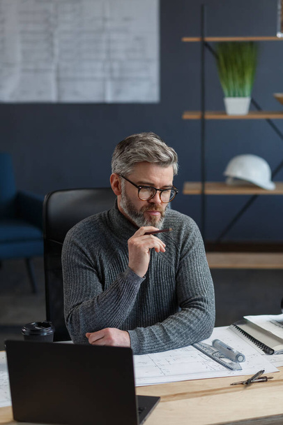 Interior designer working in office with blueprints.Engineer inspect architectural plan, sketching a construction project.Portrait of handsome bearded man at workplace. Business construction concept - 写真・画像