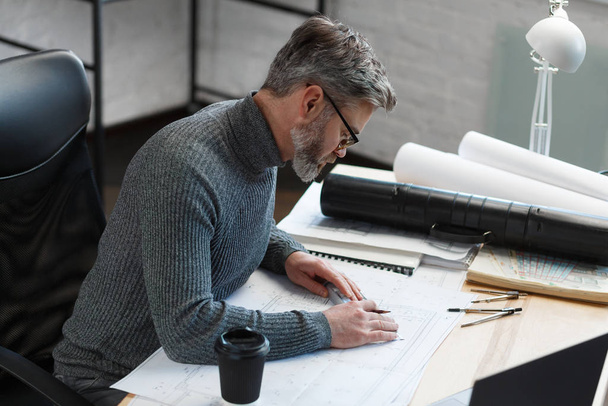 Interior designer working in office with blueprints.Engineer inspect architectural plan, sketching a construction project.Portrait of handsome bearded man at workplace. Business construction concept - Photo, image