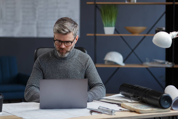 Designer working in office with laptop. Architect thinks over architectural plan, searching new ideas for construction project. Portrait of handsome bearded man sitting at workplace. Business concept - Foto, Imagem