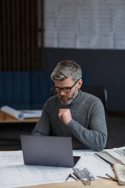 Designer working in office with laptop. Architect thinks over architectural plan, searching new ideas for construction project. Portrait of handsome bearded man sitting at workplace. Business concept - Photo, image